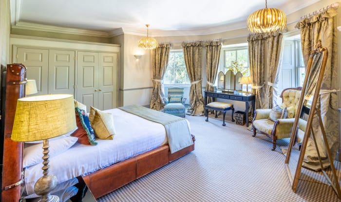 Elegant bedroom with bay windows, gold drapes, vanity, and leather‑framed bed in warm neutral tones.