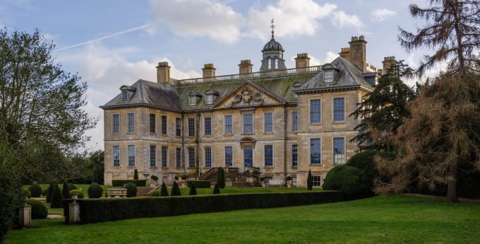 Belton House in Lincolnshire, a grand historic stone mansion surrounded by formal gardens, clipped hedges and lawns, beneath a bright, cloud-filled sky.