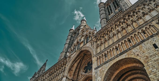 A low-angle view of a grand Gothic cathedral with intricate stonework, tall spires, and arched windows, set against a teal sky with scattered clouds.