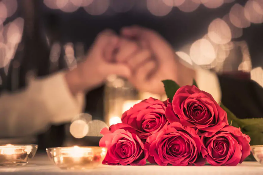 A bouquet of red roses on a candlelit table, with a blurred background of a couple holding hands, creating a romantic atmosphere with soft, warm lighting.