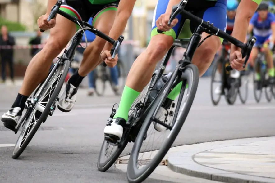 Close-up of cyclists in a road race, leaning into a turn with focus on their legs, bikes, and bright green socks, while others follow in the background.