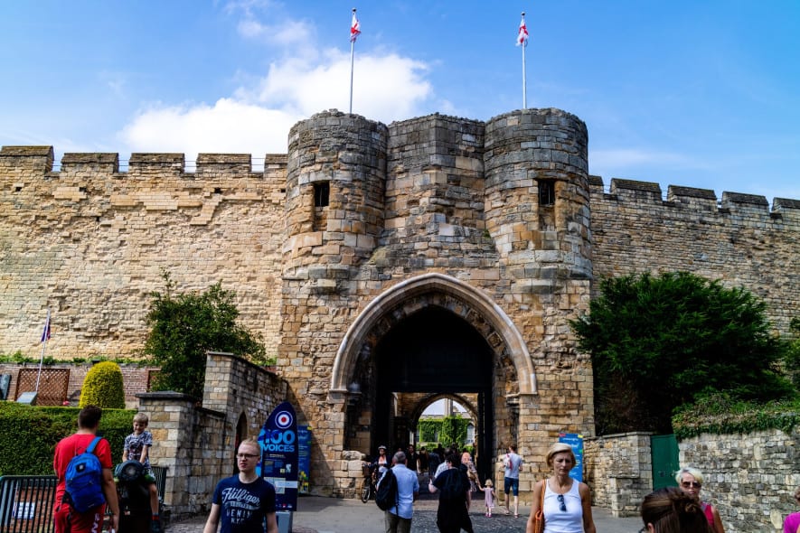 A historic stone castle gate with arched entry flanked by round towers, flying two St. George’s flags, surrounded by visitors on a sunny day with a bright blue sky.