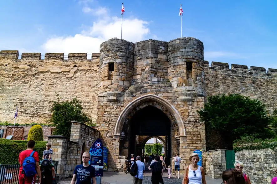 A historic stone castle gate with arched entry flanked by round towers, flying two St. George’s flags, surrounded by visitors on a sunny day with a bright blue sky.