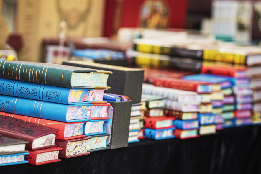 Stacks of colorful, intricately designed books displayed on a table at a market or fair, with blurred larger books and other items visible in the background.
