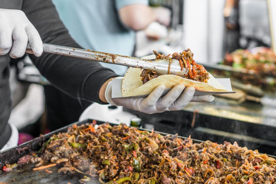 A street food vendor wearing gloves uses tongs to fill a taco with grilled meat and vegetables, with a sizzling flat-top grill piled with cooked ingredients in the background.