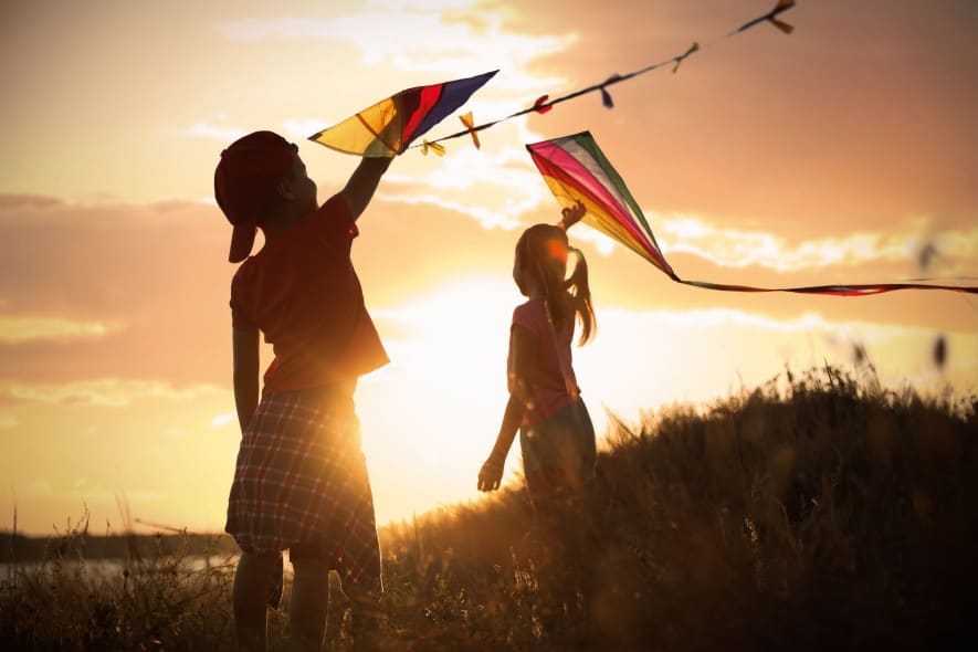 Two children flying colorful kites on a grassy hill at sunset, silhouetted against the golden sky with the sun casting a warm glow over the scene.