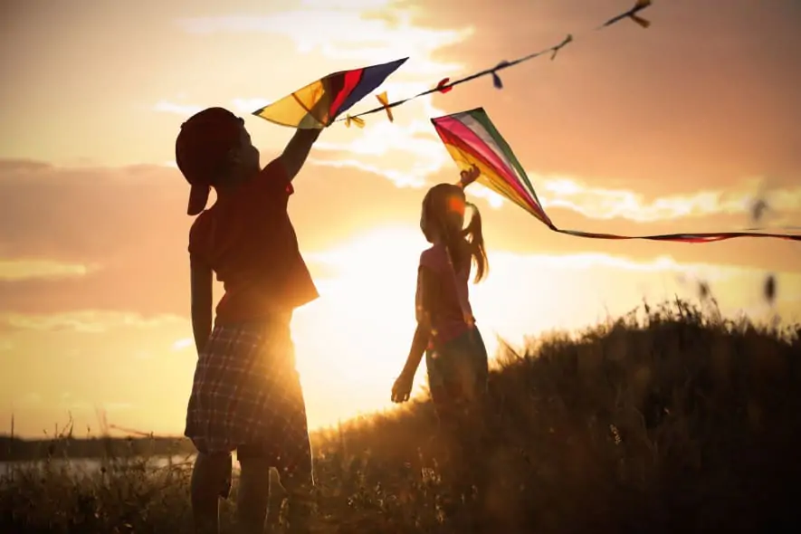 Two children flying colorful kites on a grassy hill at sunset, silhouetted against the golden sky with the sun casting a warm glow over the scene.