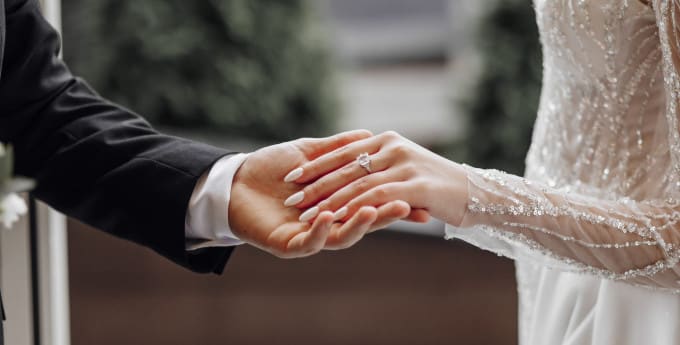 A groom in a black suit gently holds the bride’s hand, showcasing her elegant engagement ring. The bride wears a sparkling, beaded wedding gown with sheer sleeves.