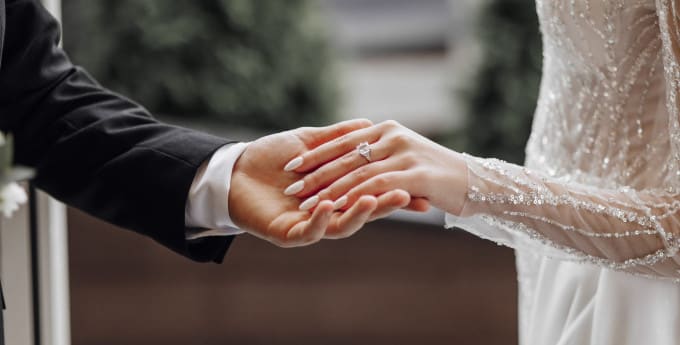 A groom in a black suit gently holds the bride’s hand, showcasing her elegant engagement ring. The bride wears a sparkling, beaded wedding gown with sheer sleeves.