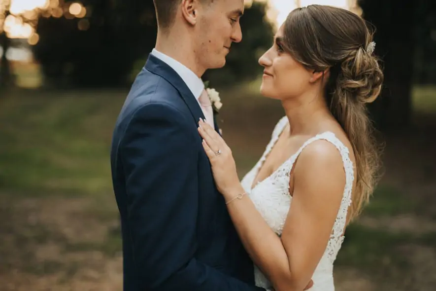 A newlywed couple gazes into each other’s eyes in an outdoor setting. The bride, in a lace wedding gown, rests her hand on the groom’s chest as he smiles warmly.