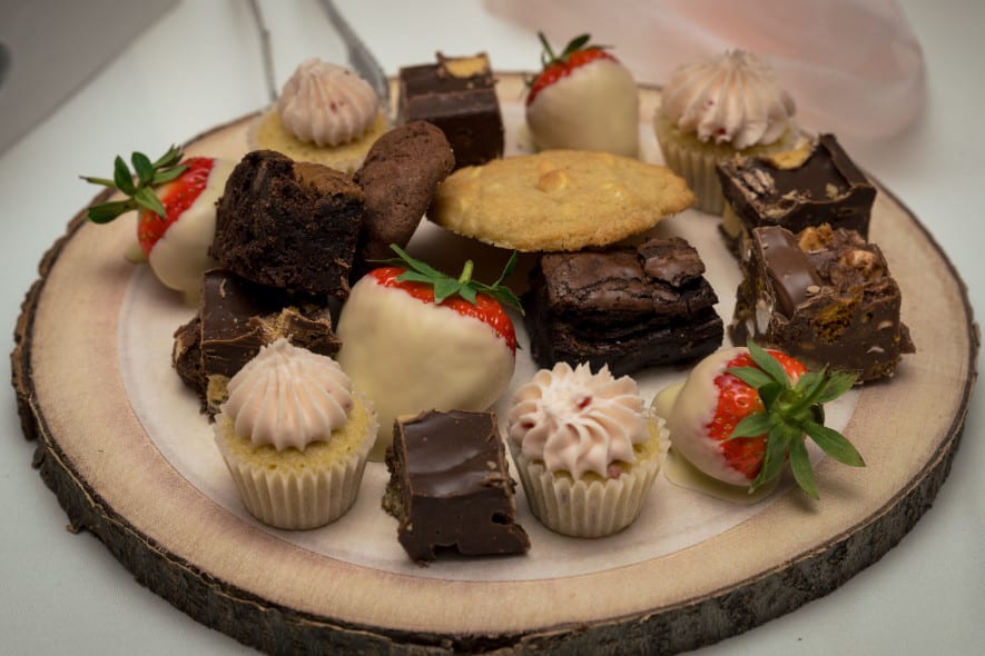 A rustic dessert platter featuring chocolate brownies, cupcakes with piped frosting, white chocolate-covered strawberries, cookies, and assorted chocolate treats.