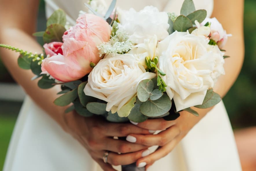 A bride in a white gown holds a bouquet of cream roses, pink peonies, and greenery. Her hands, adorned with a wedding ring and white nail polish, gently cradle the stems.