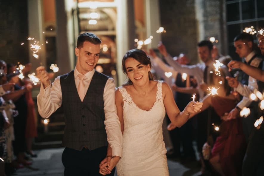 A newlywed couple walks hand in hand through a sparkler-lit send-off, smiling joyfully as friends and family celebrate their wedding night with glowing sparklers.