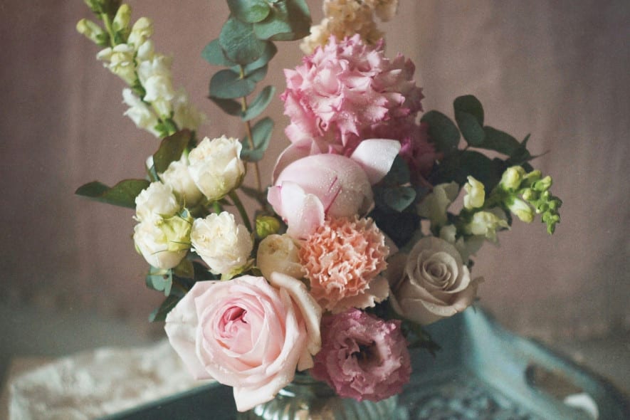 A pastel floral arrangement in a glass jar on a vintage blue tray, with lace fabric and a soft, muted background.
