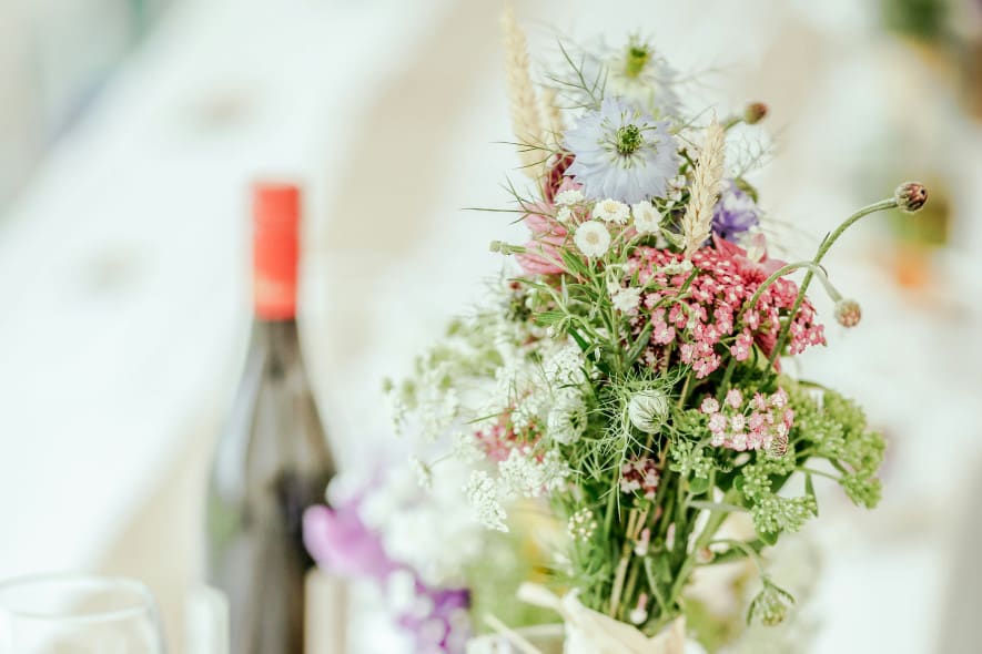 Wildflowers in pinks, yellows and blues sit in a small glass with a bottle of red wine in the background
