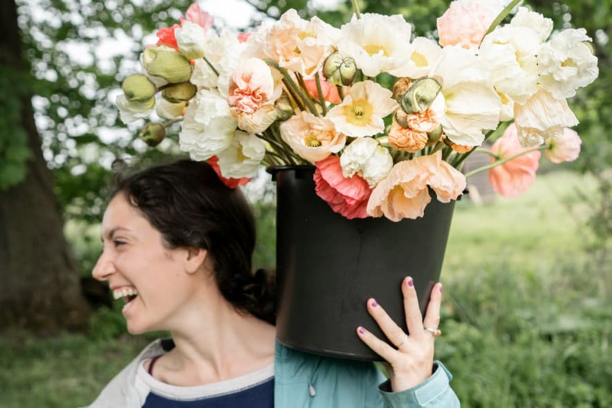 women wearing a blue coat is carrying a bucket of pastel coloured flowers in a black bucket, and resting this on her shoulder as she looks left and is laughing