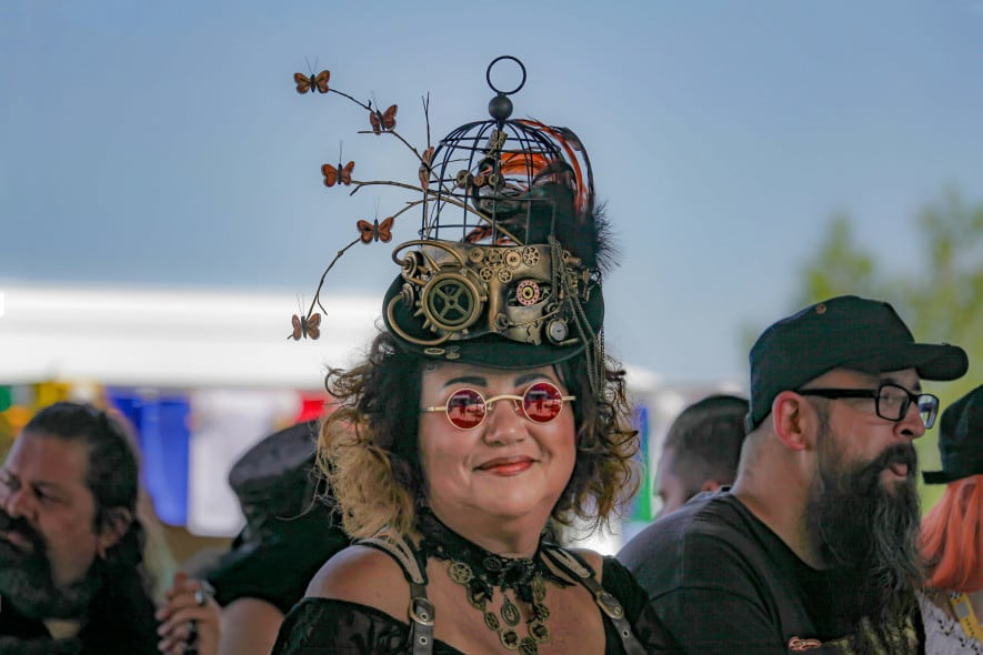 women wears a steampunk style hat, with metal cogs and metal butterflies attached, while smiling at the camera and wearing round sunglasses with a pink lens
