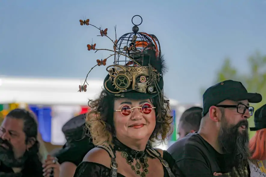 women wears a steampunk style hat, with metal cogs and metal butterflies attached, while smiling at the camera and wearing round sunglasses with a pink lens
