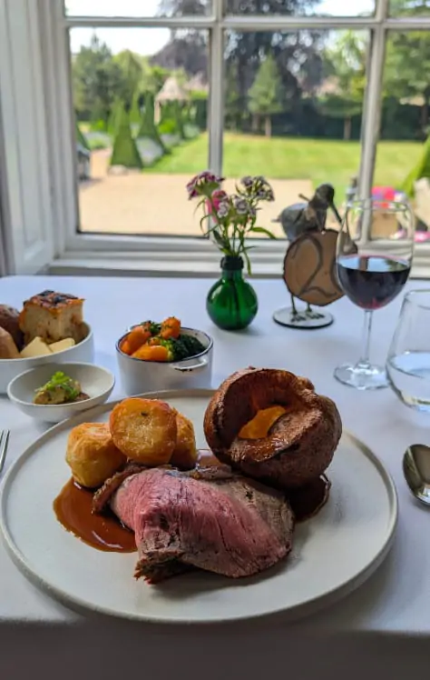 A traditional British roast dinner with beef, Yorkshire pudding, roast potatoes, vegetables, and red wine, served at a window table with garden view.