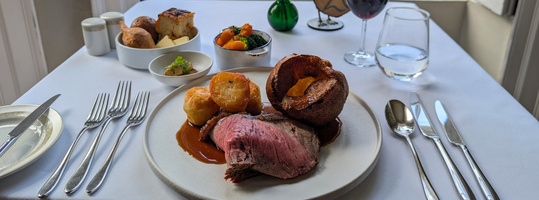 A traditional British roast dinner with beef, Yorkshire pudding, roast potatoes, vegetables, and red wine, served at a window table with garden view.