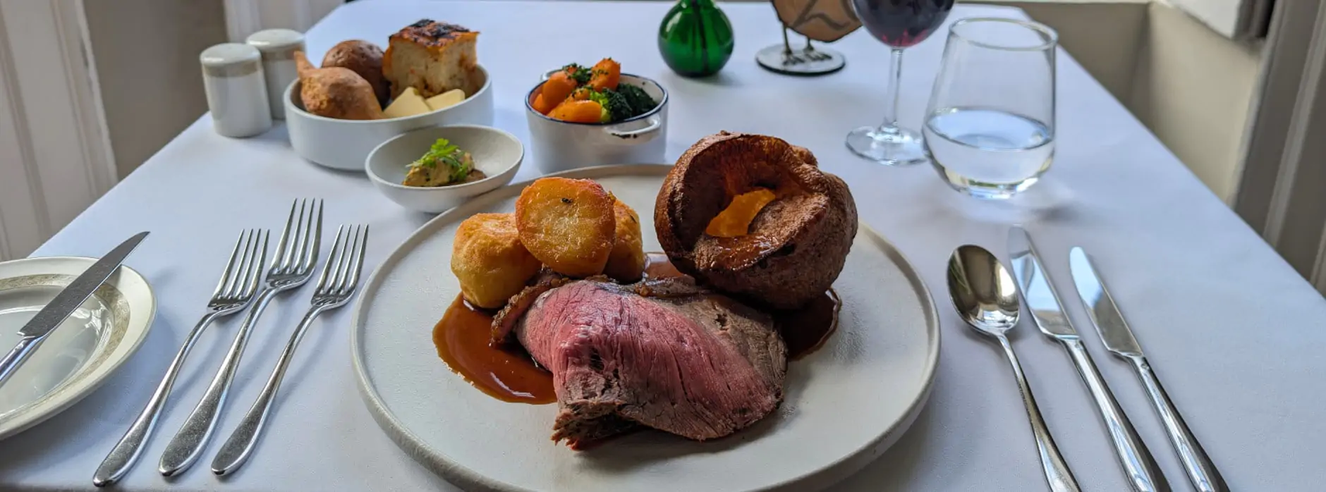 A traditional British roast dinner with beef, Yorkshire pudding, roast potatoes, vegetables, and red wine, served at a window table with garden view.