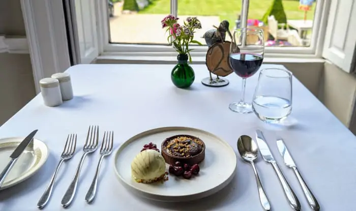Chocolate tart with ice cream and cherries on a white plate, served at a window-side table with wine and a view of topiary garden.