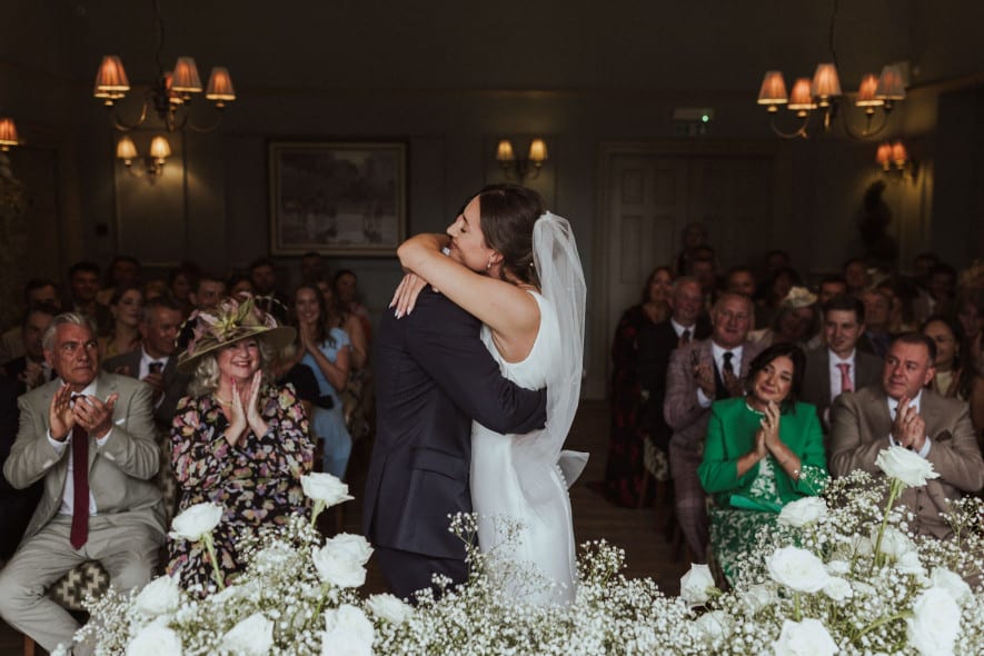 Bride and groom share a joyful embrace during their indoor ceremony at Washingborough Hall, surrounded by guests and white florals.