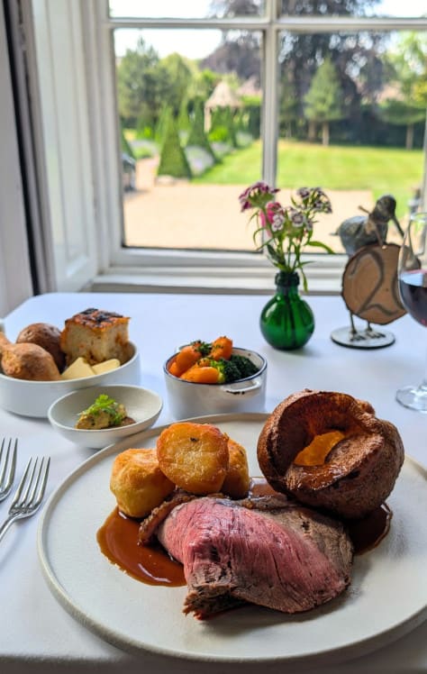 Elegant Sunday roast dinner with beef, roast potatoes, Yorkshire pudding, vegetables, and wine, served by a window overlooking a garden.