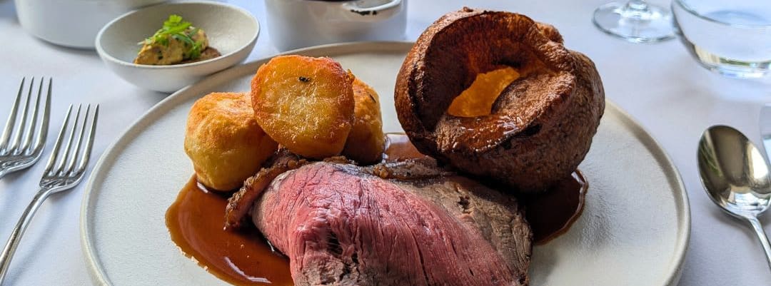 Elegant Sunday roast dinner with beef, roast potatoes, Yorkshire pudding, vegetables, and wine, served by a window overlooking a garden.