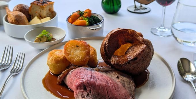 Elegant Sunday roast dinner with beef, roast potatoes, Yorkshire pudding, vegetables, and wine, served by a window overlooking a garden.