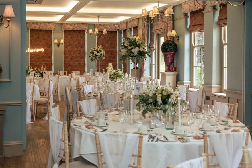 Elegant wedding reception setup with round tables, white linens, floral centrepieces, and natural light in a refined, pastel-toned room.