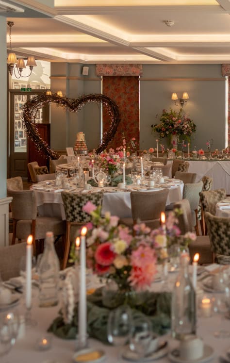 Romantic wedding reception with floral centrepieces, candles, elegant round tables, and a heart-shaped light feature in a refined venue.