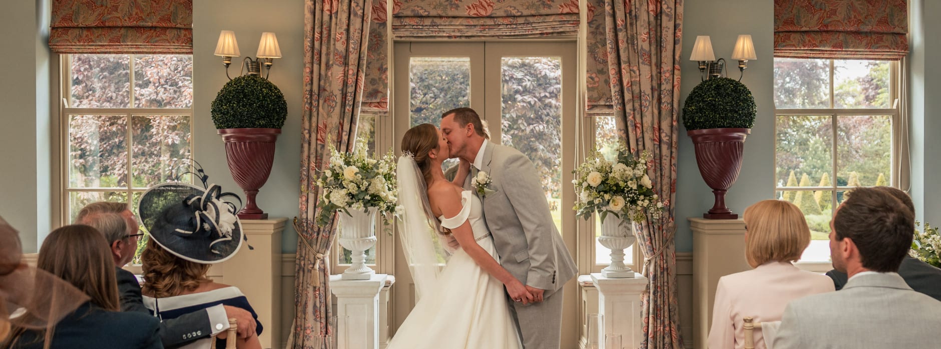 Bride and groom share their first kiss during the wedding ceremony, surrounded by guests, flowers, candles, and elegant décor.