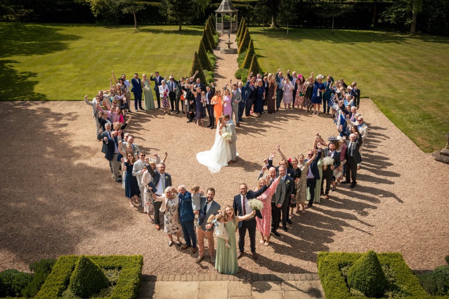 Wedding guests form a large heart shape around the newlywed couple kissing in the centre of a garden courtyard on a sunny day.