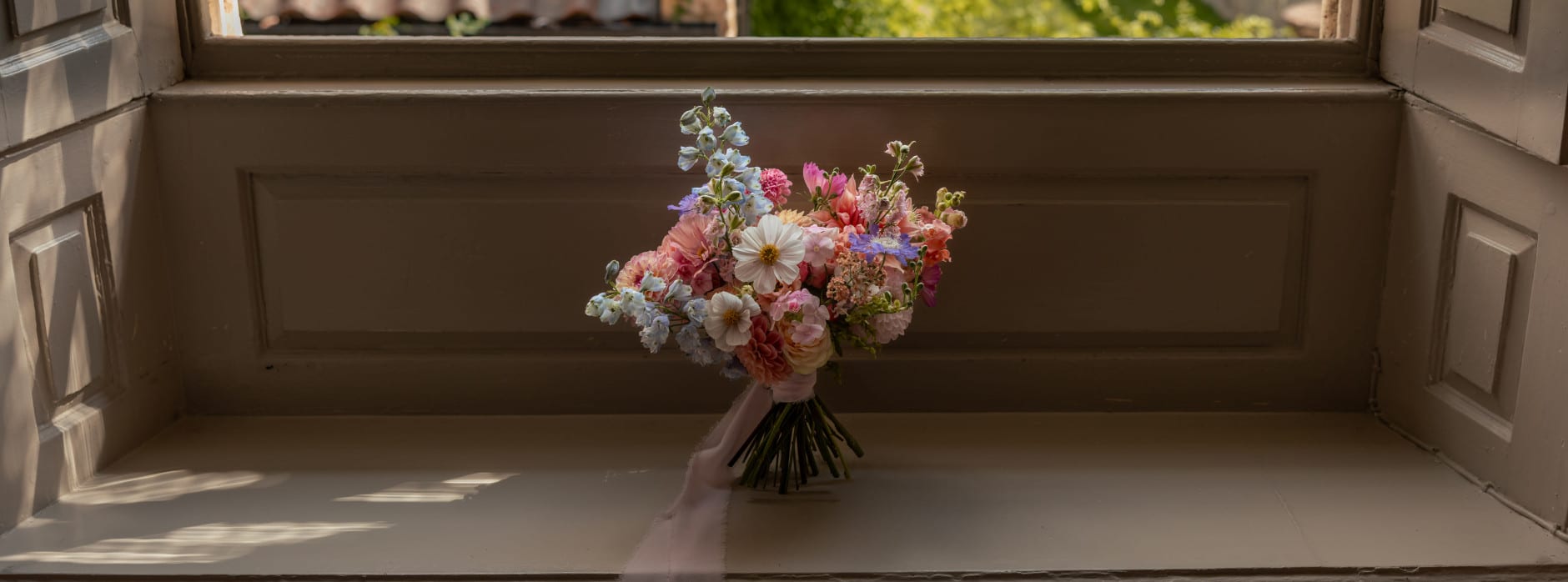 A pastel wedding bouquet with pink, white, and blue flowers rests on a windowsill with soft light streaming through.