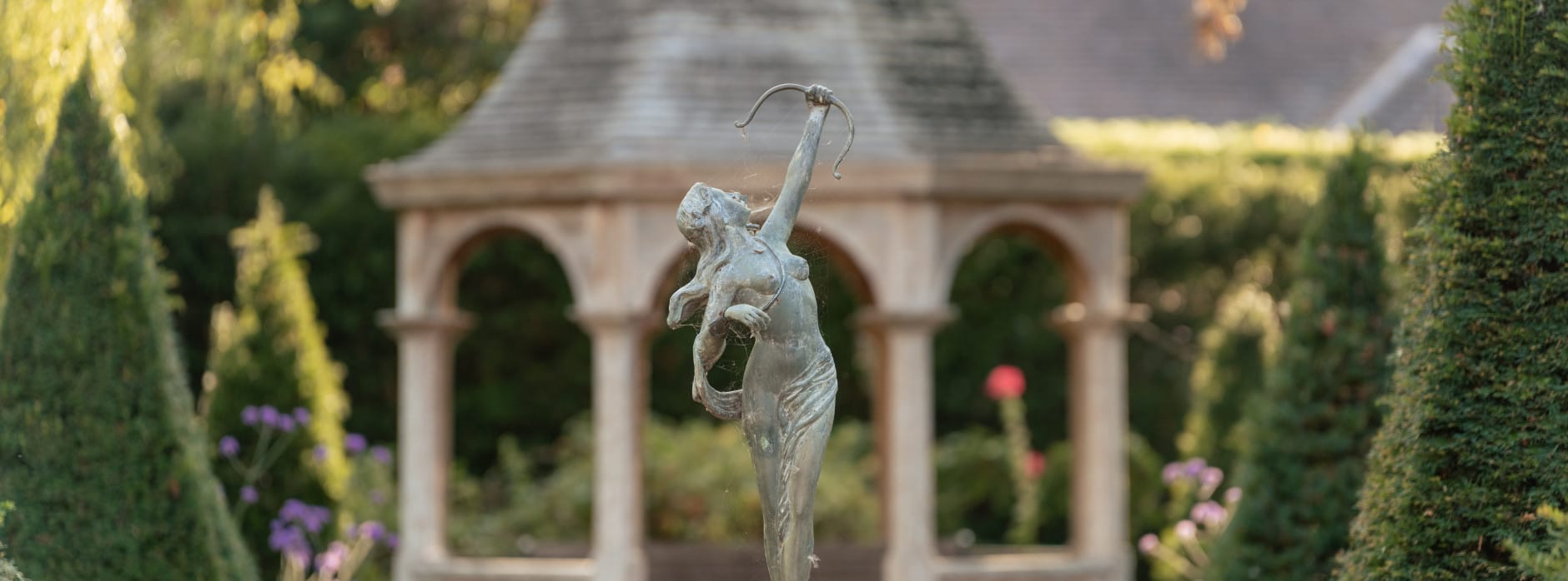 A bronze statue of a woman with a bow stands on a fountain, framed by topiary and a gazebo in the background.
