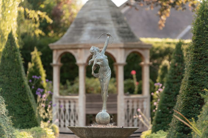 A bronze statue of a woman with a bow stands on a fountain, framed by topiary and a gazebo in the background.