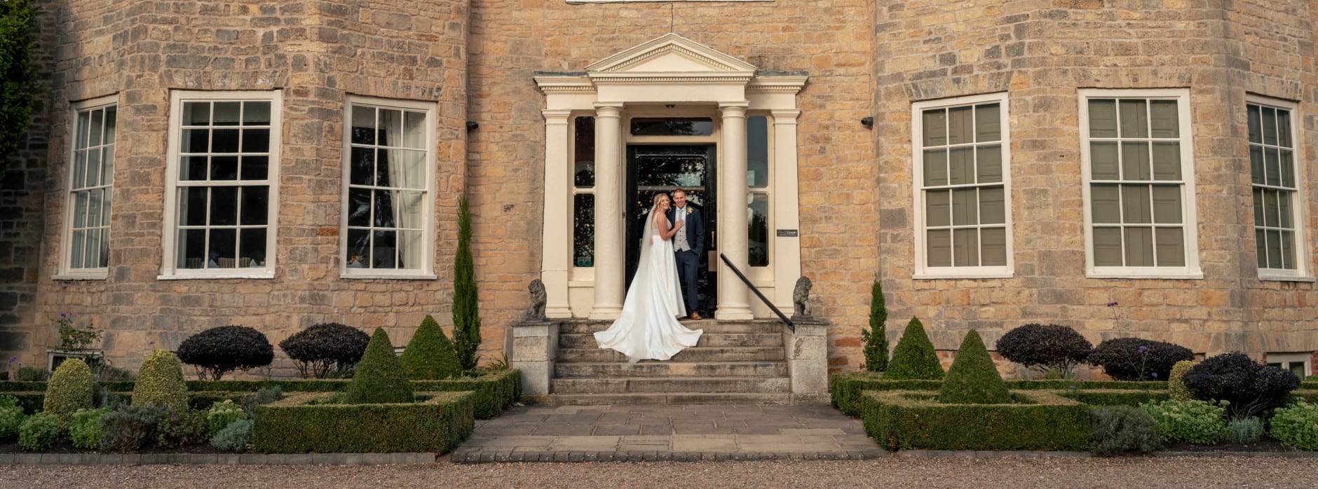 Bride and groom stand in front of a grand stone manor house entrance, smiling together on the steps beneath elegant columns.