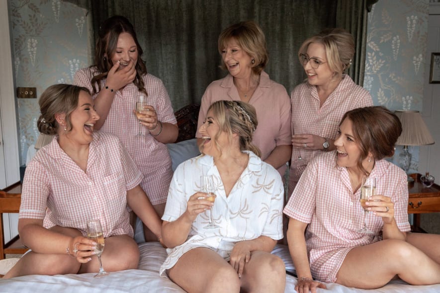 Bride, bridesmaids, and her mother laugh together on a bed in matching pyjamas, holding glasses of champagne.