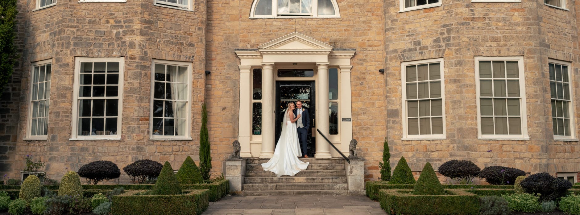 Bride and groom stand in front of a grand stone manor house entrance, smiling together on the steps beneath elegant columns.