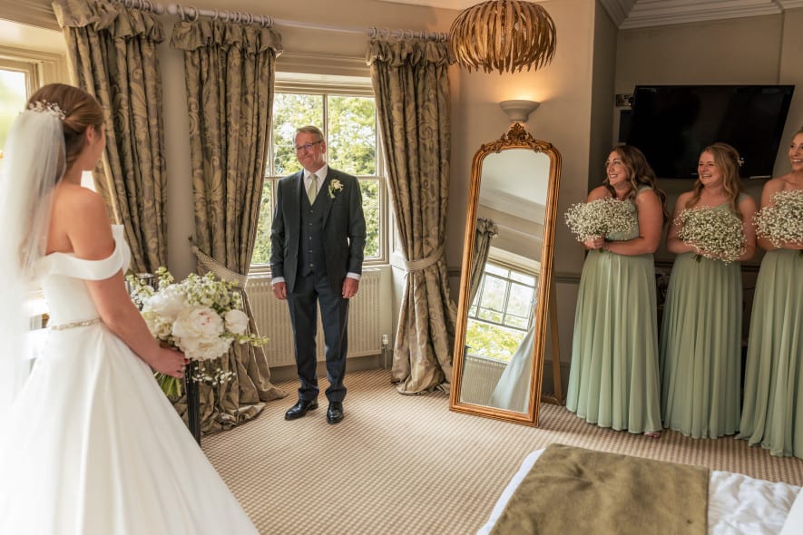 Bride shares an emotional first look with her father in a decorated room, while bridesmaids in sage dresses smile holding bouquets.