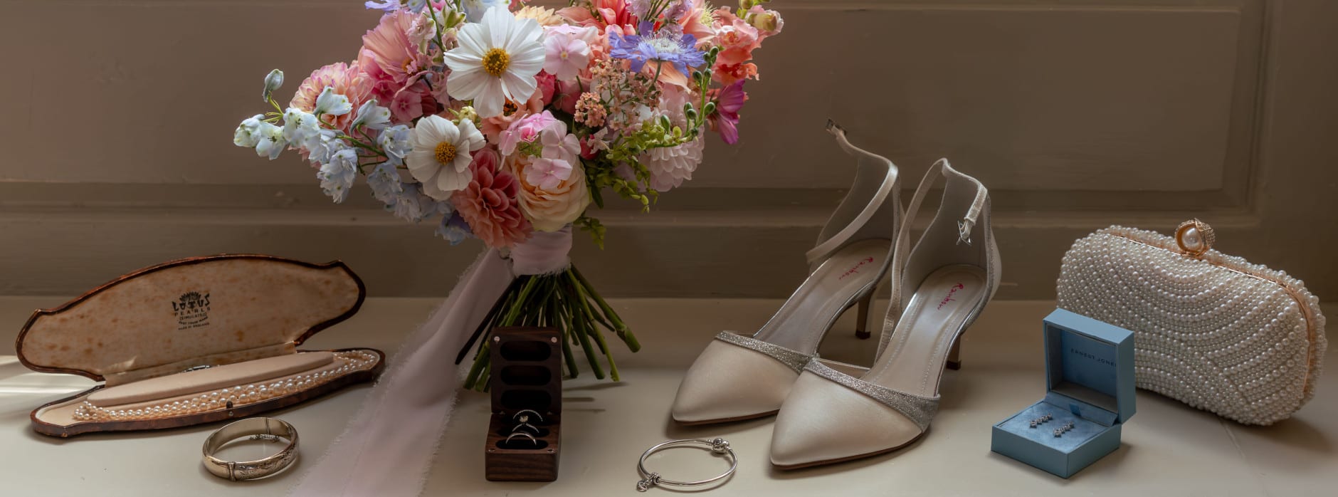 Bridal accessories including bouquet, pearl necklace, rings, silver bracelet, ivory heels, earrings, and pearl clutch displayed on a windowsill.